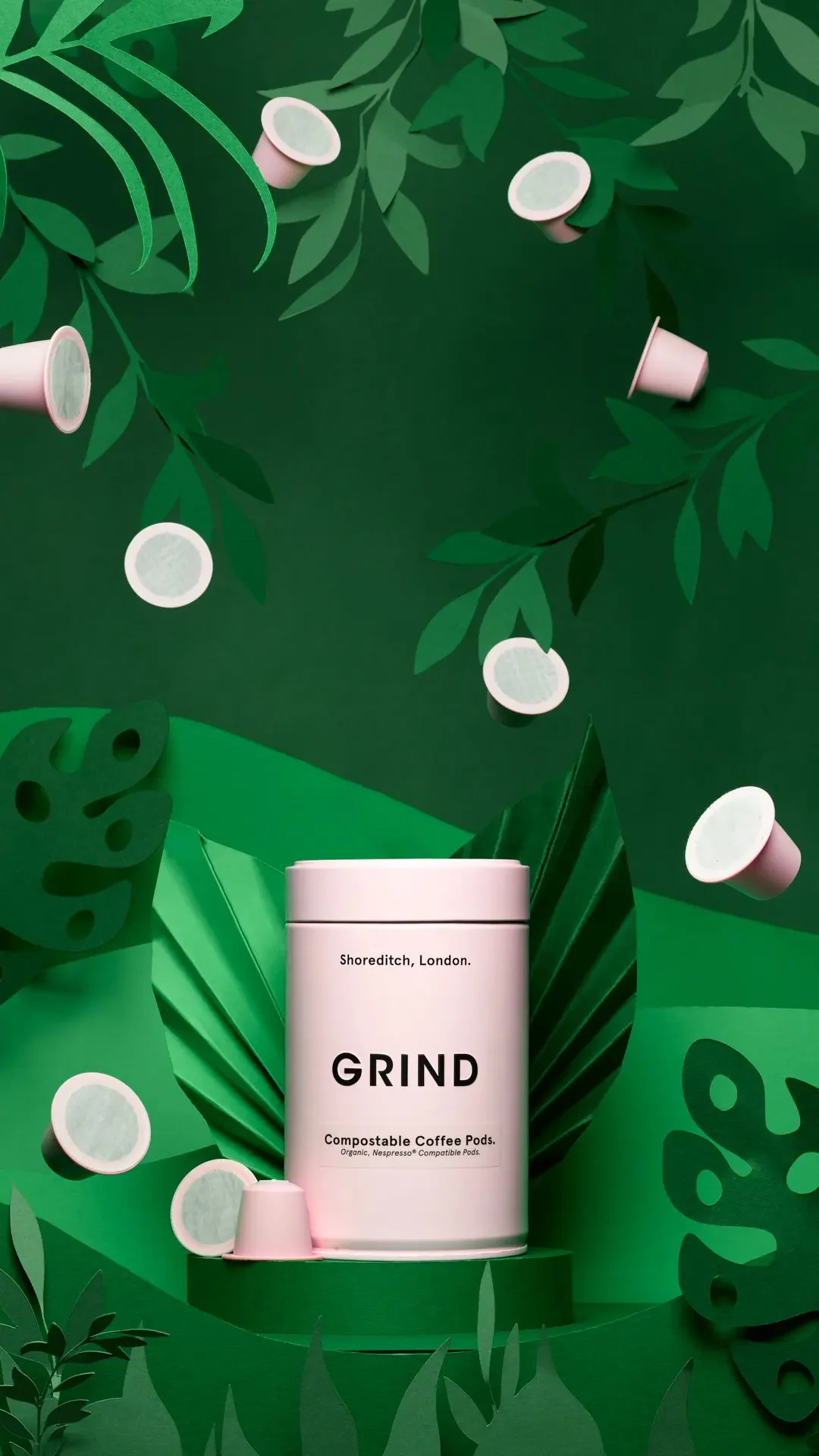 A pink GRIND coffee-pod tin and pods among layered green paper leaves.