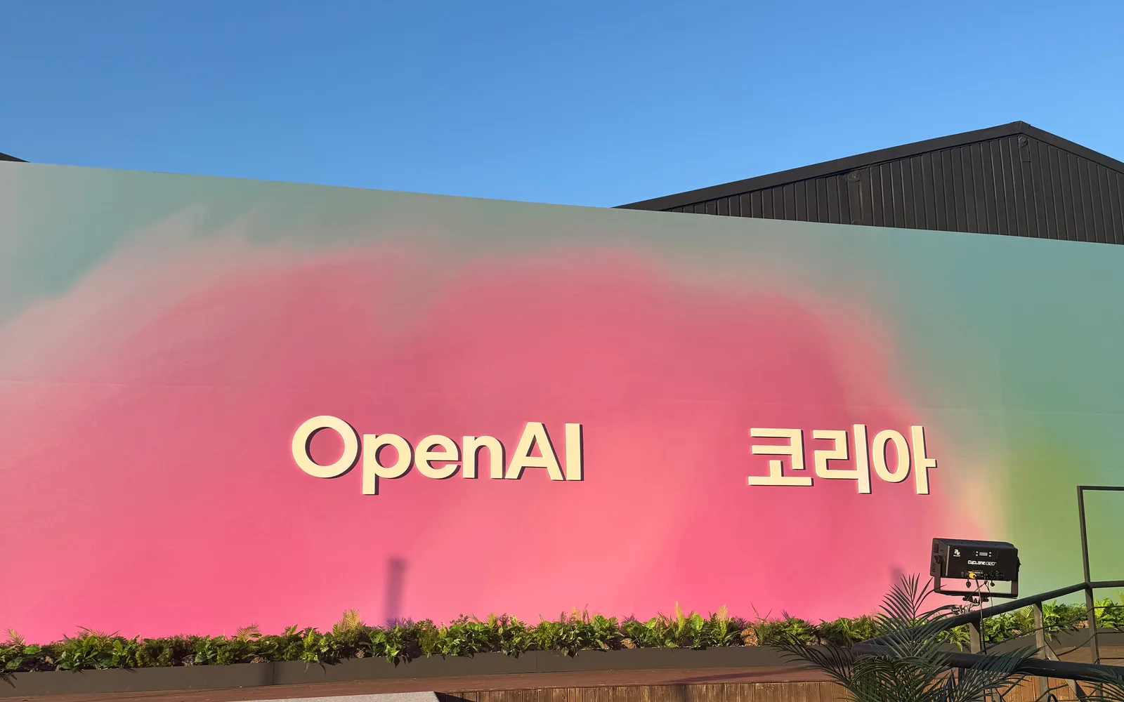 The OpenAI 코리아 sign on a pink and green wall outside the launch venue in Seoul, under a clear blue sky.